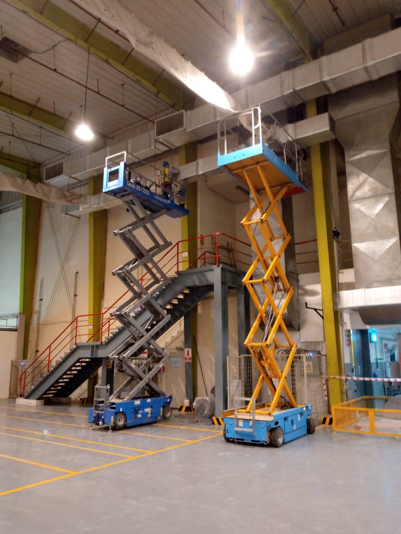 Two scissor lifts in an industrial facility, one fully extended with a worker platform and the other partially raised, showcasing their functionality for elevated work.